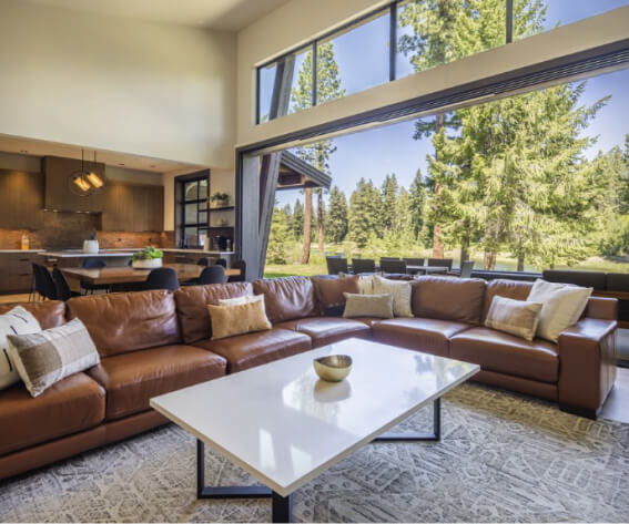 Spacious living room at Suncadia Resort with a large leather sectional sofa, white coffee table, and a view of tall trees through wide windows. Dining area and kitchen visible in the background, epitomizing the charm of Suncadia real estate.